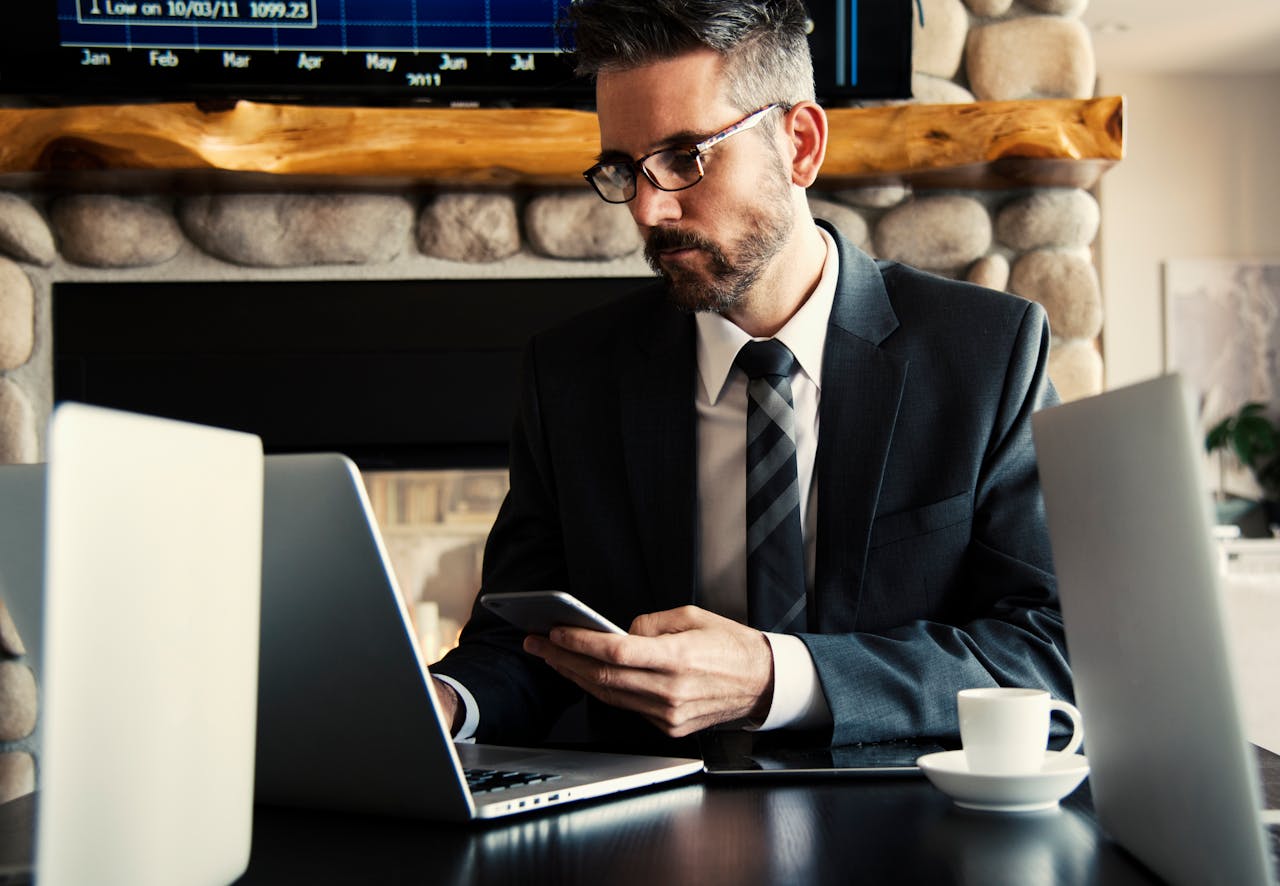 Businessman in formal attire working on a laptop and smartphone at an indoor office desk.